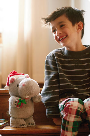 Young boy smiling and sitting next to a stuffed hippo wearing a Santa hat and holding a gift.