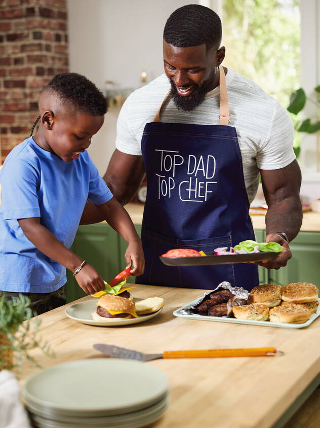 Father wearing Top Dad' apron in kitchen with young son plating food.