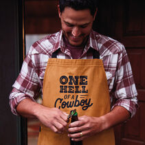 Man opening a bottle wearing a grill apron that reads “One hell of a cowboy”
