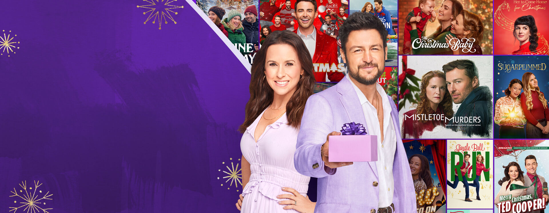 Lacey Chabert and Tyler Hines standing in front of Hallmark holiday movie posters