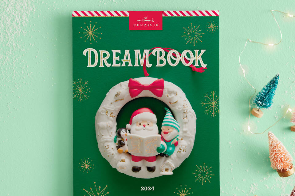 The 2024 Dreambook cover is green with a candycane stripe along the top. It features an ornament of Santa reading to a penguin and gnome.