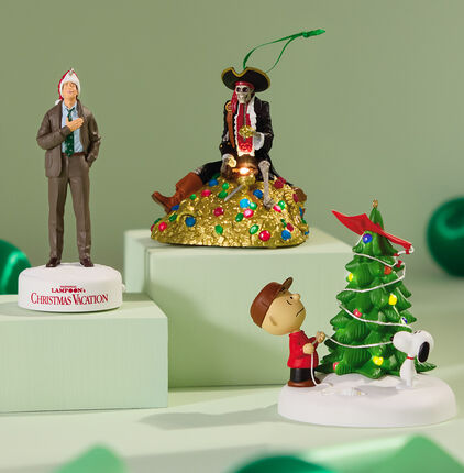 A Christmas Vacation ornament, Pirates of the Caribbean ornament, and Charlie Brown and Snoopy ornament on a mint background. 