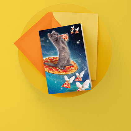Greeting card featuring kitten in space standing on a pizza while eating a slice. Additional pizza slices with feather wings fly around them.