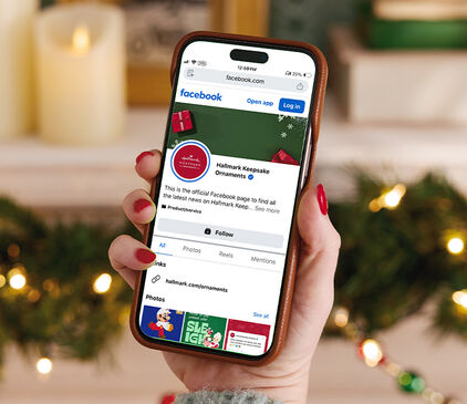 Womans hand holding a smartphone open to the Hallmark Keepsake Ornaments Facebook page.