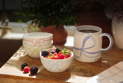 Ceramic mug and set of 3 bowls with pastel ribbons on table with fruit