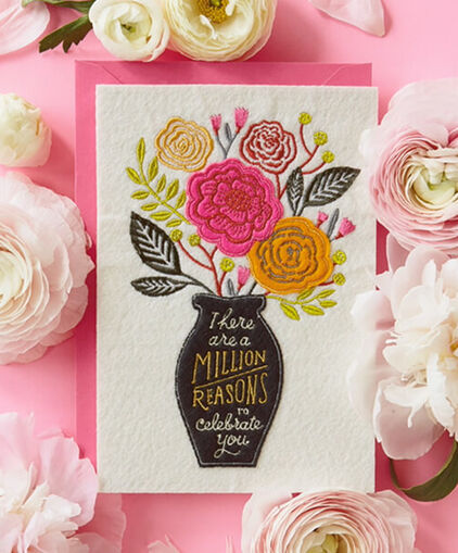 Embroidered felt card surrounded by white and pink flowers. 