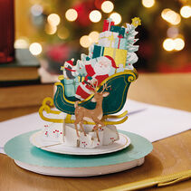 Musical pop-up Christmas greeting card with die-cut Santa Claus, penguin and reindeer in a jade green sleigh filled with gifts and a Christmas tree with foil accents with laser-cut details
