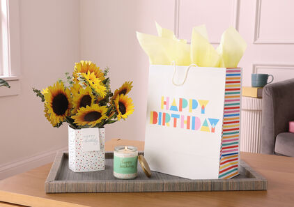 Paper bouquet, candle and gift bag that all say ‘Happy Birthday’ on a coffee table