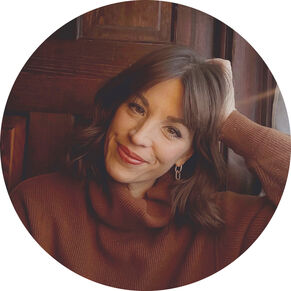 Woman with brown hair and a brown cowl-neck sweater leaning her head on her hand and smiling