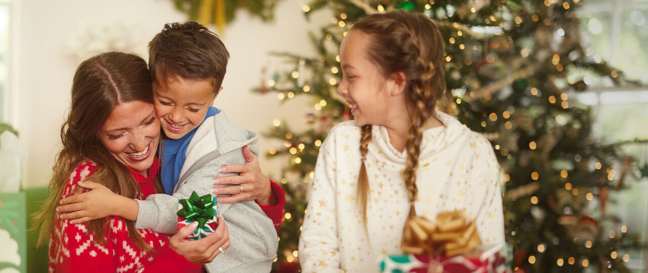 Woman hugging a young boy with a young girl smiling at them in front of a Christmas tree.