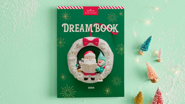 The 2024 Dreambook cover is green with a candycane stripe along the top. It features an ornament of Santa reading to a penguin and gnome.